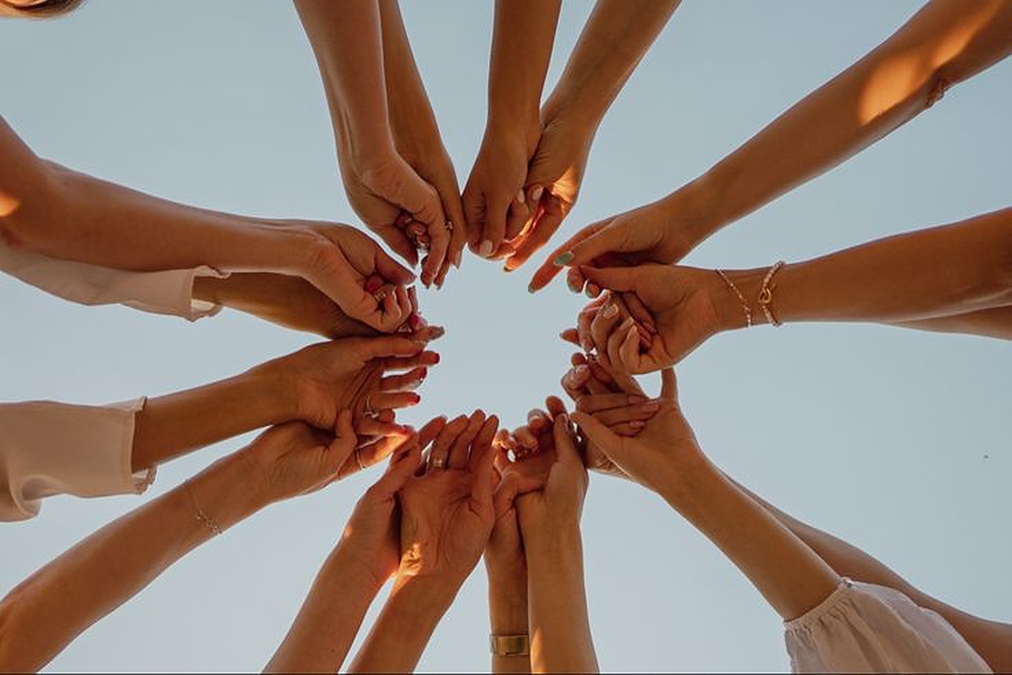 Mãos de mulheres que se encontram em círculo, vistas de baixo, sob um céu claro